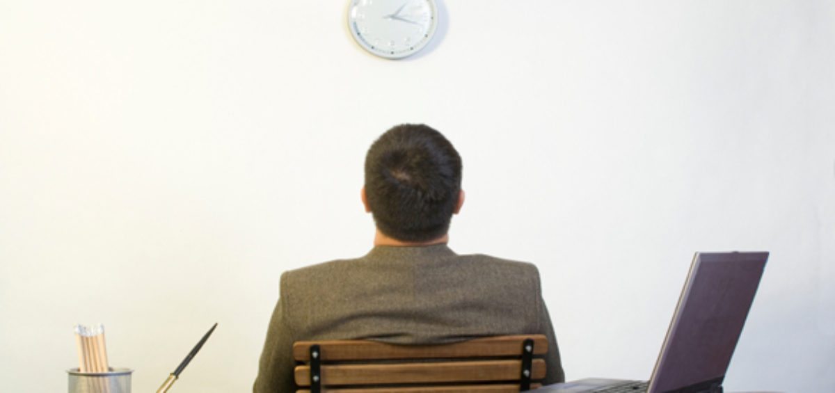 Man in office staring at clock (clock-watching), showing lack of care for his work; an indication of low employee engagement